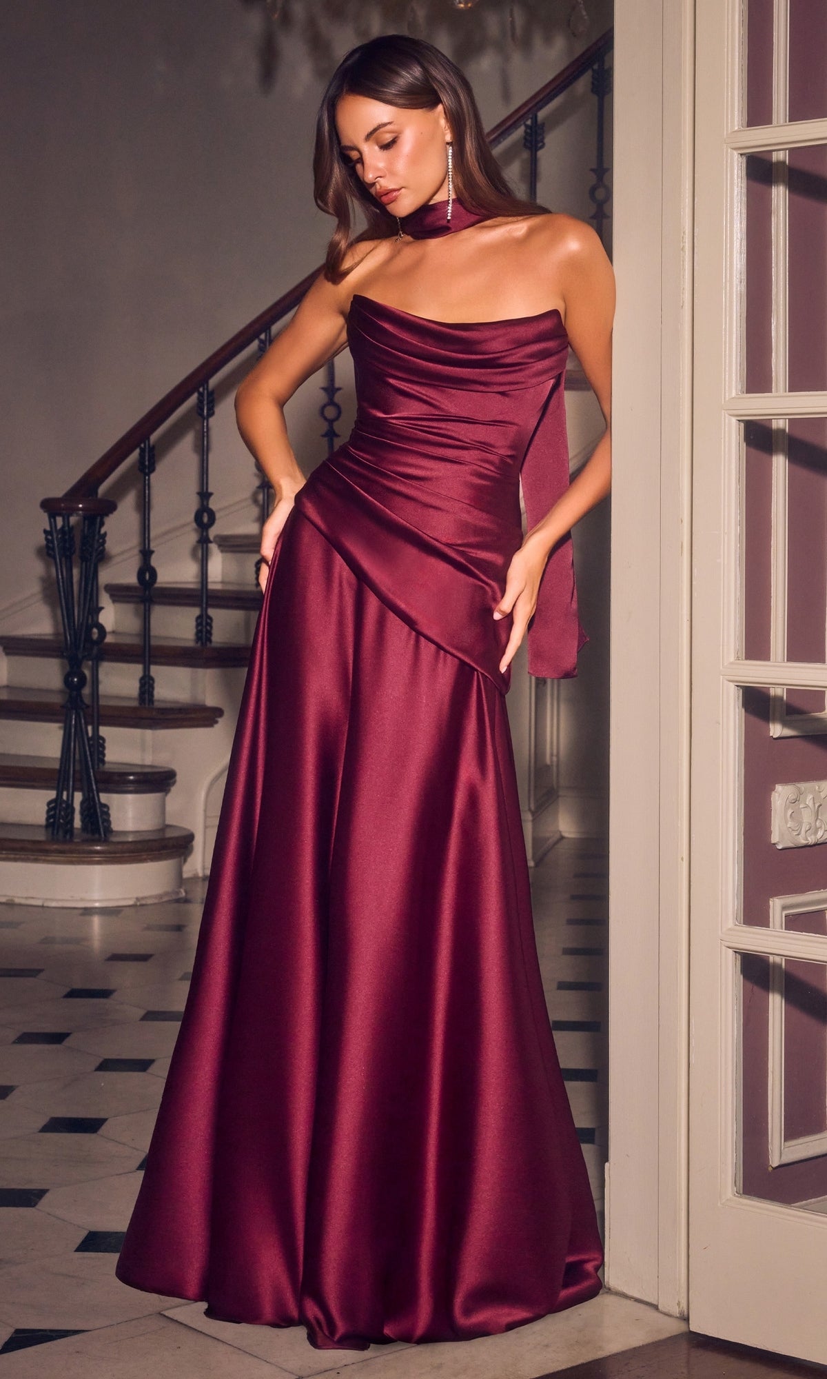 A woman stands indoors by a staircase wearing the Ladivine 7432 long prom dress in burgundy, featuring a floor-length satin gown with a ruched bodice, asymmetrical neckline, matching choker, and an elegant lace-up back for added detail and sophistication.